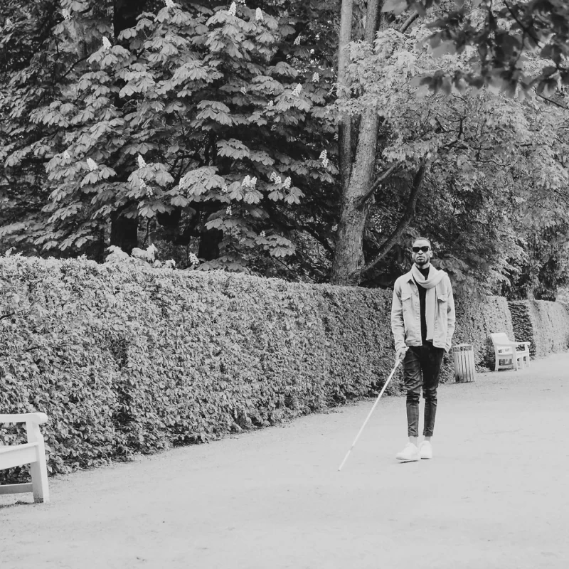 A blind man with a white cane standing in a park next to a nature.